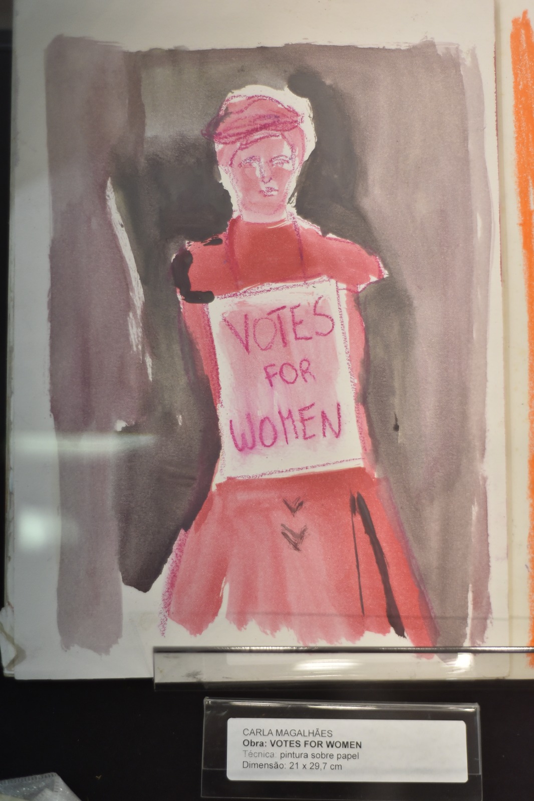 Foto de uma pintura em aquarela, mostrando uma figura feminina em tons de rosa e vermelho. Há um cartaz pendurado em seu pescoço, no qual se lê "VOTES FOR WOMEN", que pode ser traduzido como "votem em mulheres"