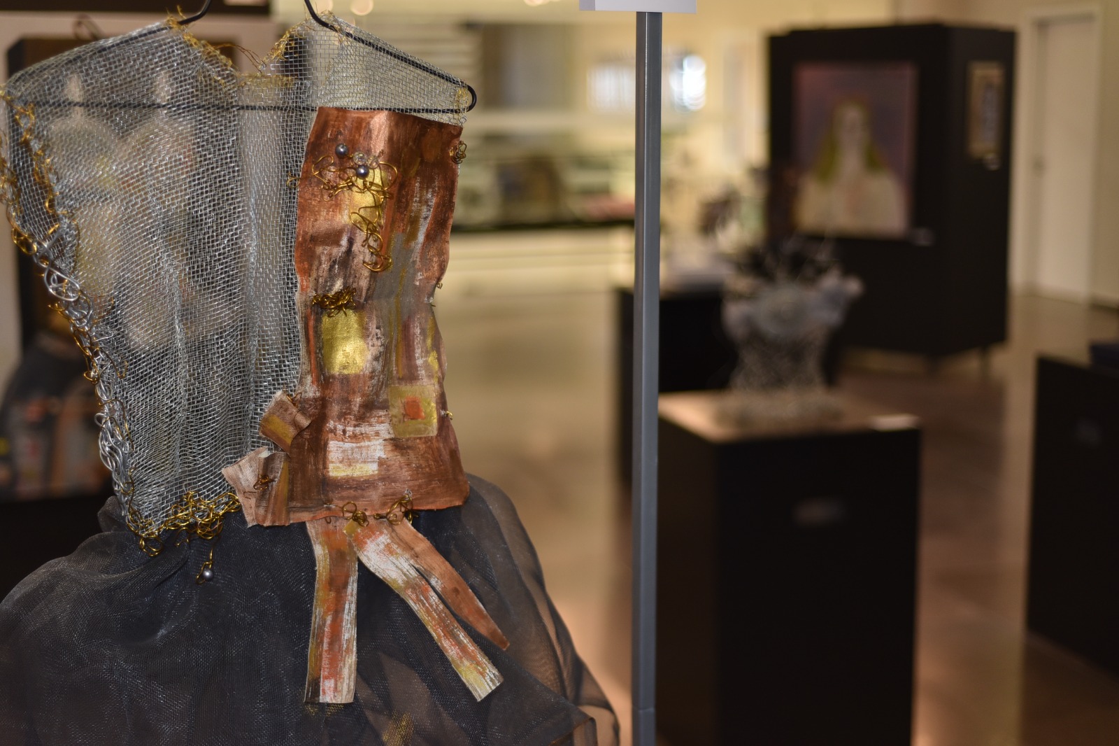 Foto em plano fechado de uma escultura em forma de corselete, feita com fios metálicos prateado e dourados e tecido pintado em cores terrosas.