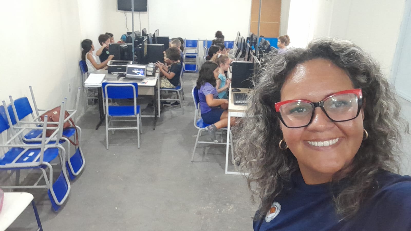 Selfie da professora Themis sorrindo, com a sala de informática ao fundo sendo usada por crianças. Ela é uma mulher parda, com cabelos grisalhos, longos e crespos, usa óculos e uma camiseta azul marinho.