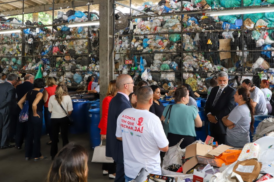 Foto feita durante a visita do presidente do TST, Lelio Bentes, ao Centro de Educação Ambiental, em Porto Alegre/RS, no dia 26 de fevereiro. Ele veste terno e tem cabelos grisalhos e barba branca. Está prestando atenção a uma catadora, que veste uniforme cinza e tem os cabelos castanhos, longos e presos. Há pessoas prestando atenção na conversa deles e outras pessoas espalhadas na estação de triagem, visitantes e trabalhadores. Ao fundo, uma grade que vai até o teto está cheia de sacos de lixo.