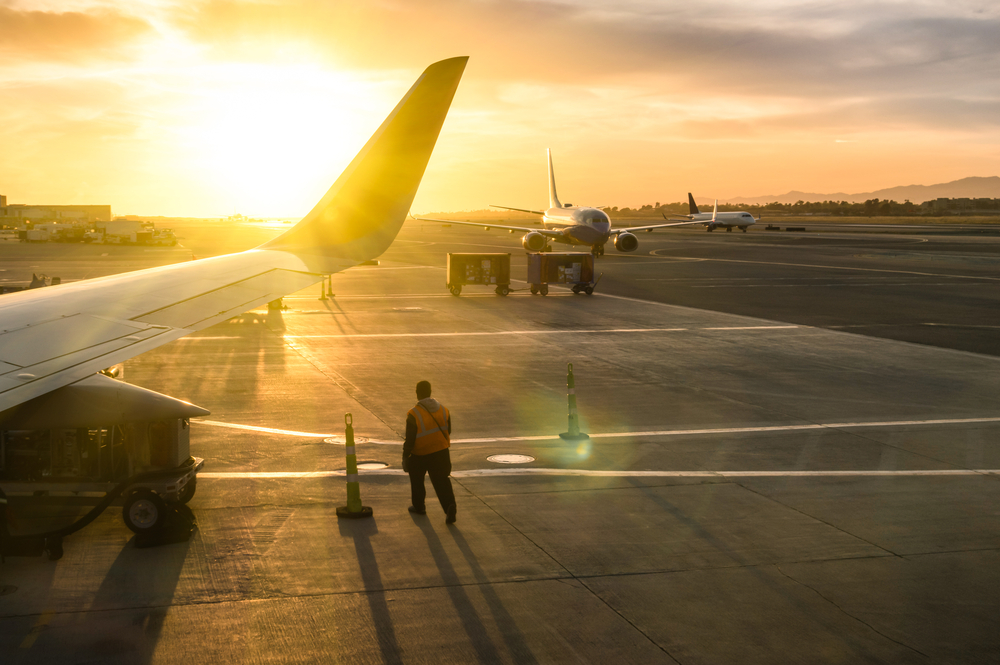 trabalhador em terminal de aeroporto, próximo a aviões. Ao fundo, há o pôr do sol.