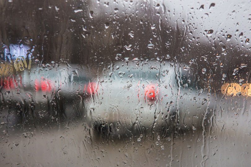 Foto ilustrativa de um carro no trânsito, com muita chuva. Água escorre pelo vidro pára-brisa.