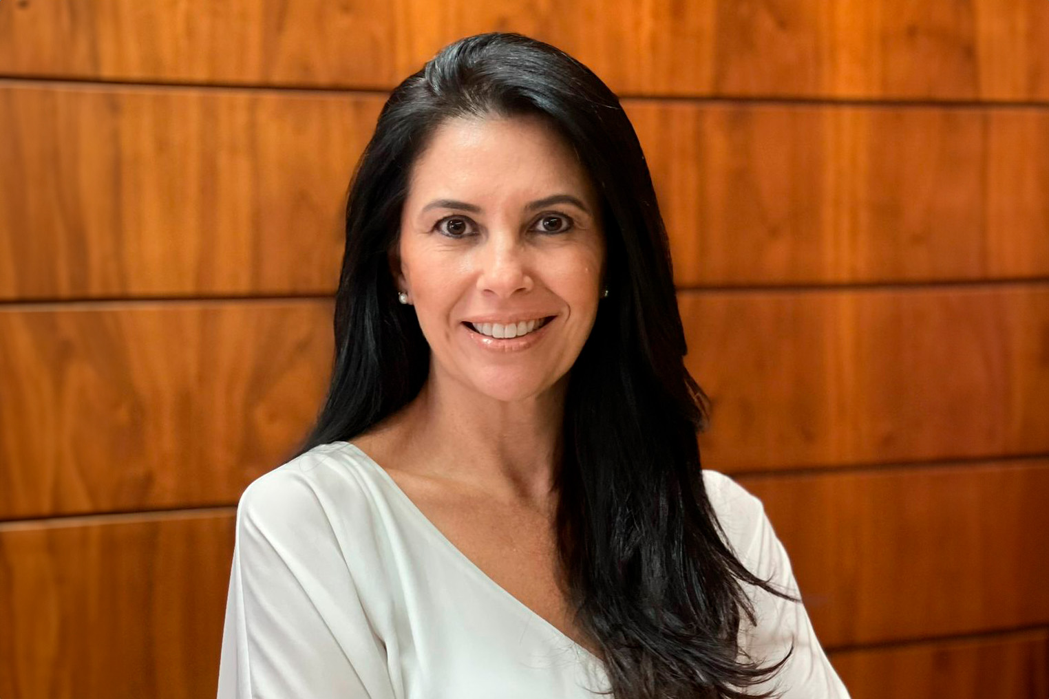 Retrato da juíza Aline Fagundes. Ela é branca, tem cabelo liso e preto comprido. Usa uma blusa branca ,está de braços cruzados e sorri. Ao fundo aparece uma parede de madeira.