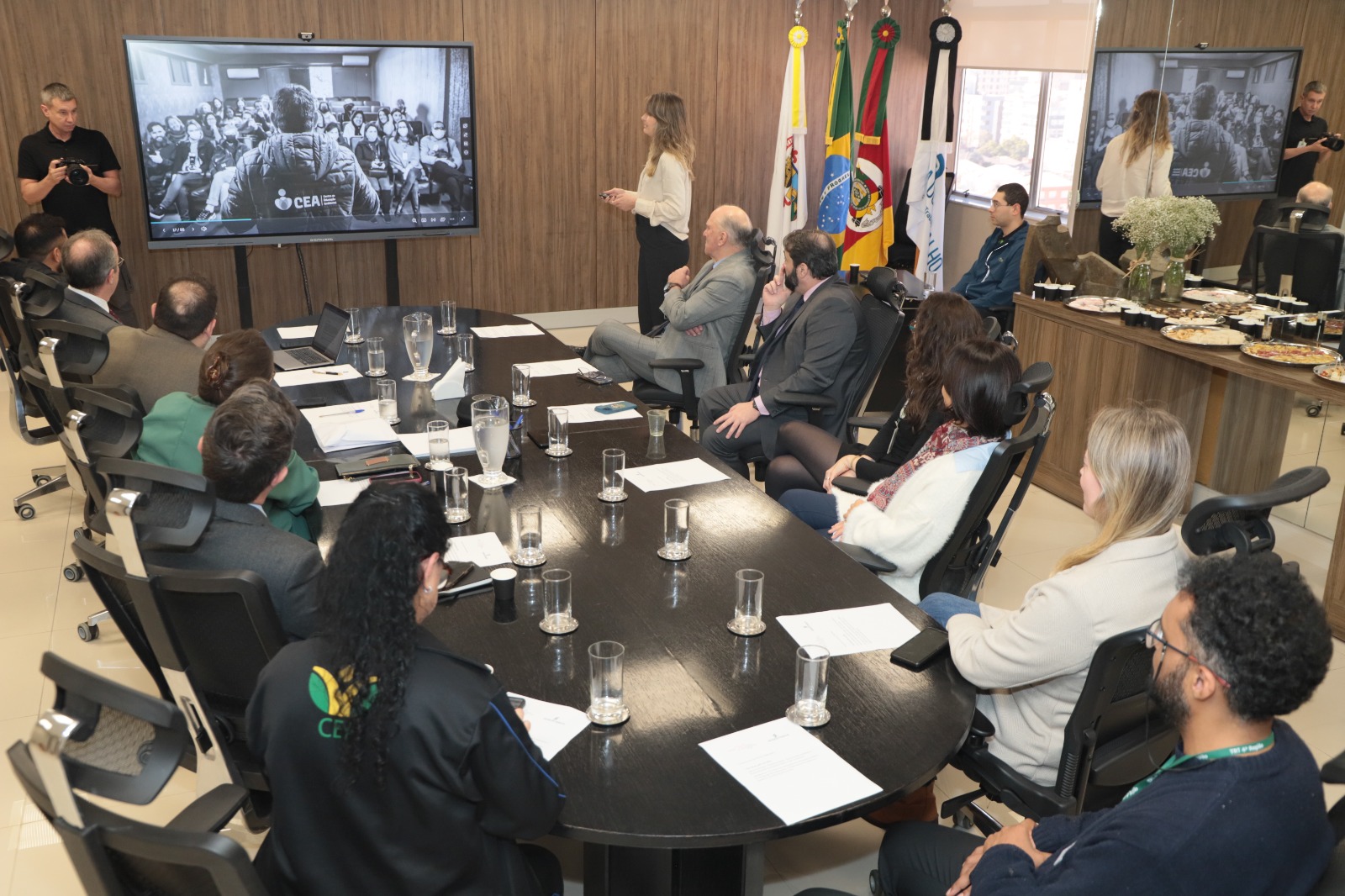 A imagem mostra uma reuniãono Salão Nobre da Presidência. As pessoas estão sentadas em torno de uma mesa grande e oval, equipada com copos de água, jarros de vidro, papéis e laptops. Na parede ao fundo, há bandeiras oficiais. Anita está de pé ao lado de uma tela grande, apresentando algo para os participantes sentados. Na tela, há uma imagem em preto e branco de uma reunião no CEA, mostrando uma pessoa de costas falando para um grupo.  A atmosfera parece ser profissional e formal.