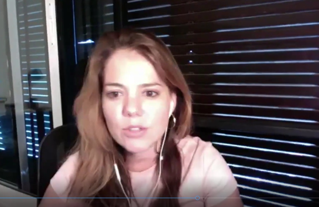 A imagem mostra Mayra, uma mulher com cabelo castanho claro, solto, usando fones de ouvido brancos. Ela veste uma camiseta clara e está em um ambiente fechado, com persianas ao fundo e uma janela de vidro à esquerda.