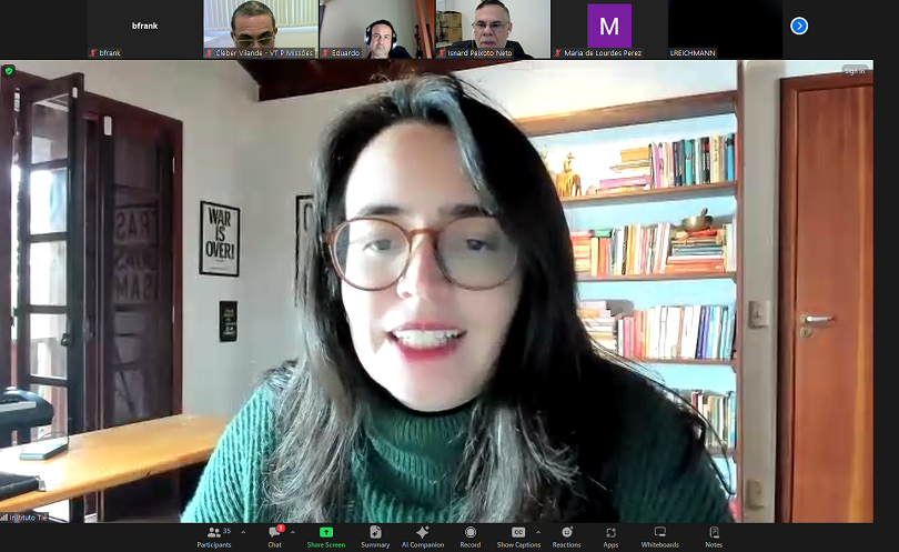 Uma mulher com óculos e suéter verde fala em uma videoconferência do Zoom. Ao fundo, uma estante com livros e pôsteres na parede. Há vários participantes na chamada, mostrados na parte superior da tela.