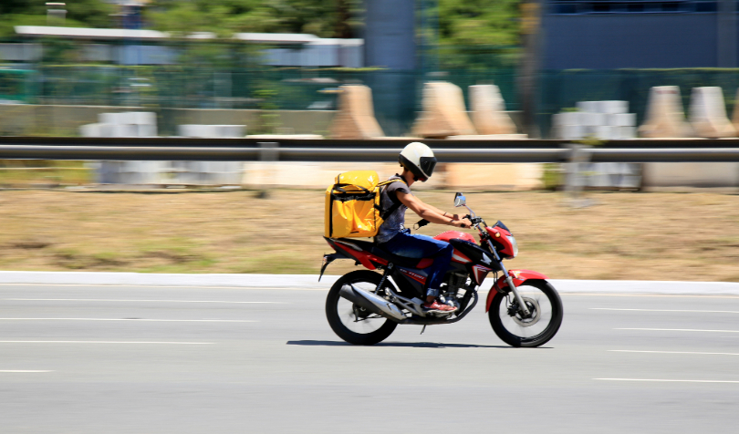  Motoboy dirigindo na cidade. A moto é vermelha, com uma caixa amarela na parte de trás