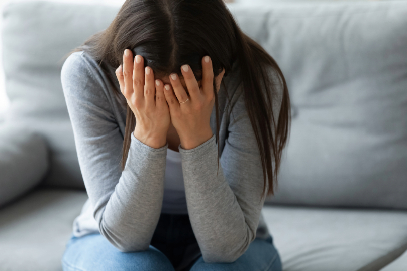 Mulher triste. Ela tem a pele clara e longos cabelos castanhos. Ela cobre o rosto com as mãos.