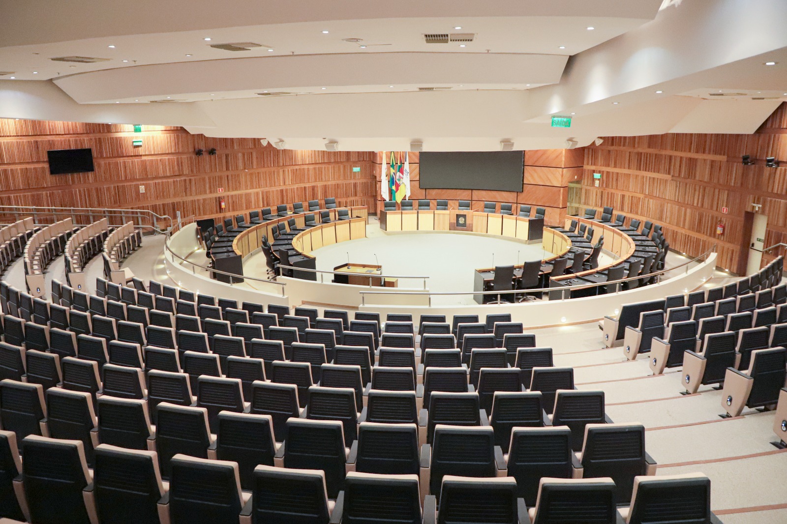 Foto do Plenário do TRT-RS vazio, após os reparos. Em primeiro plano aparecem as cadeiras da plateia, e ao fundo a nave central, com as cadeiras dos desembargadores, bandeiras e um telão desligado ao centro.