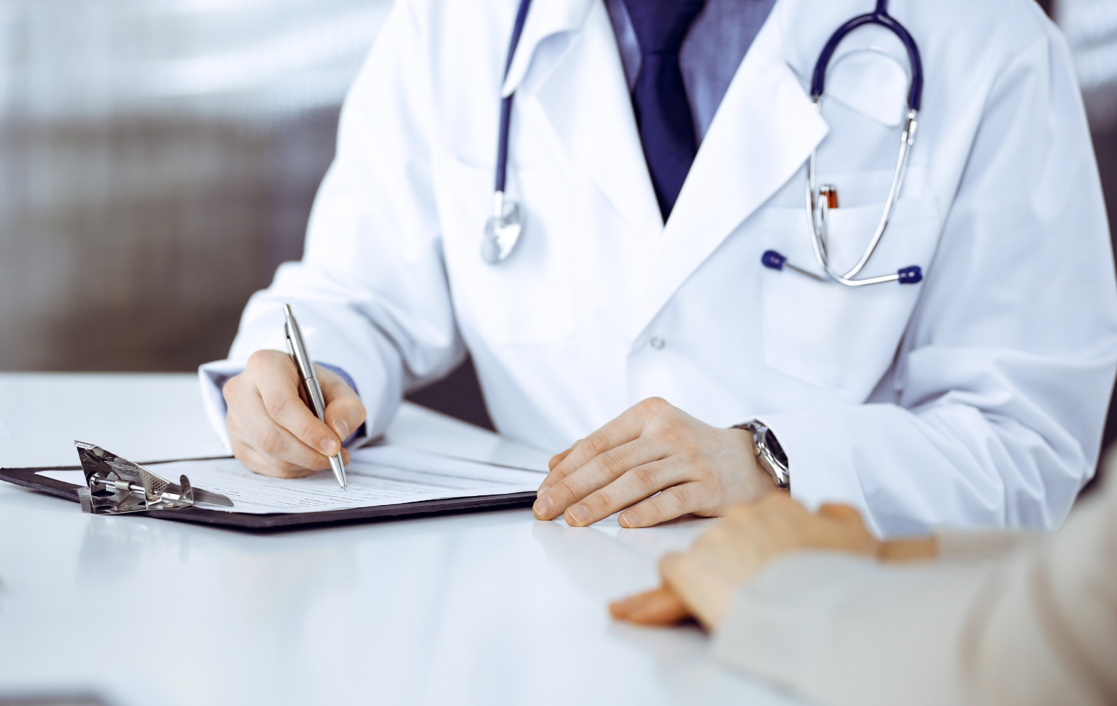 Foto de um médico com um estetoscópio no pescoço atendendo uma pessia e escrevendo algo num papel_.jfif