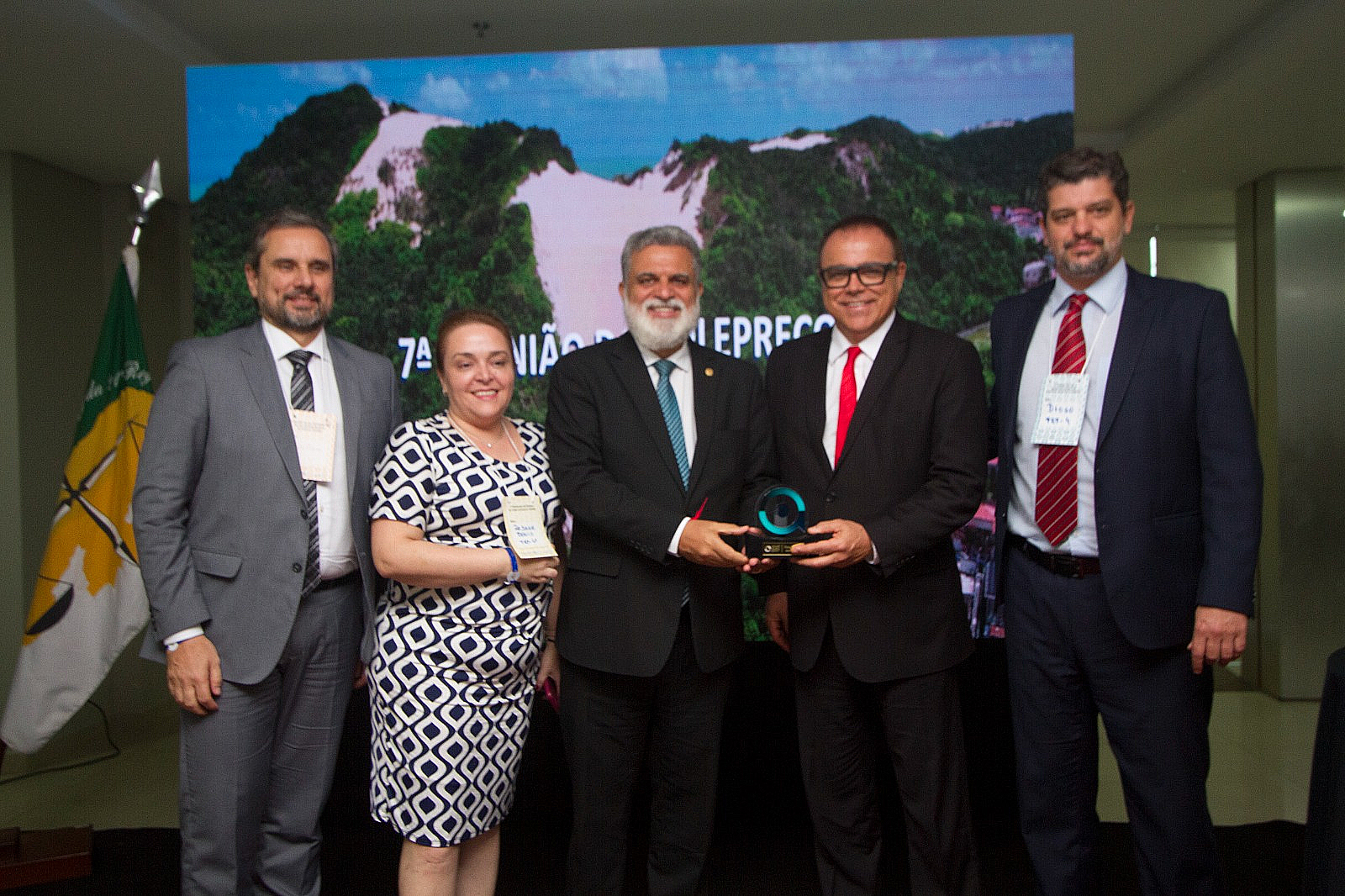 Adolfo, Rejane, Lelio, Geraldo e Diogo posam para a foto. Lelio e Geraldo estão ao centro segurando um trofeu. Ao fundo o telão mostra uma foto de dunas de areia em Natal.