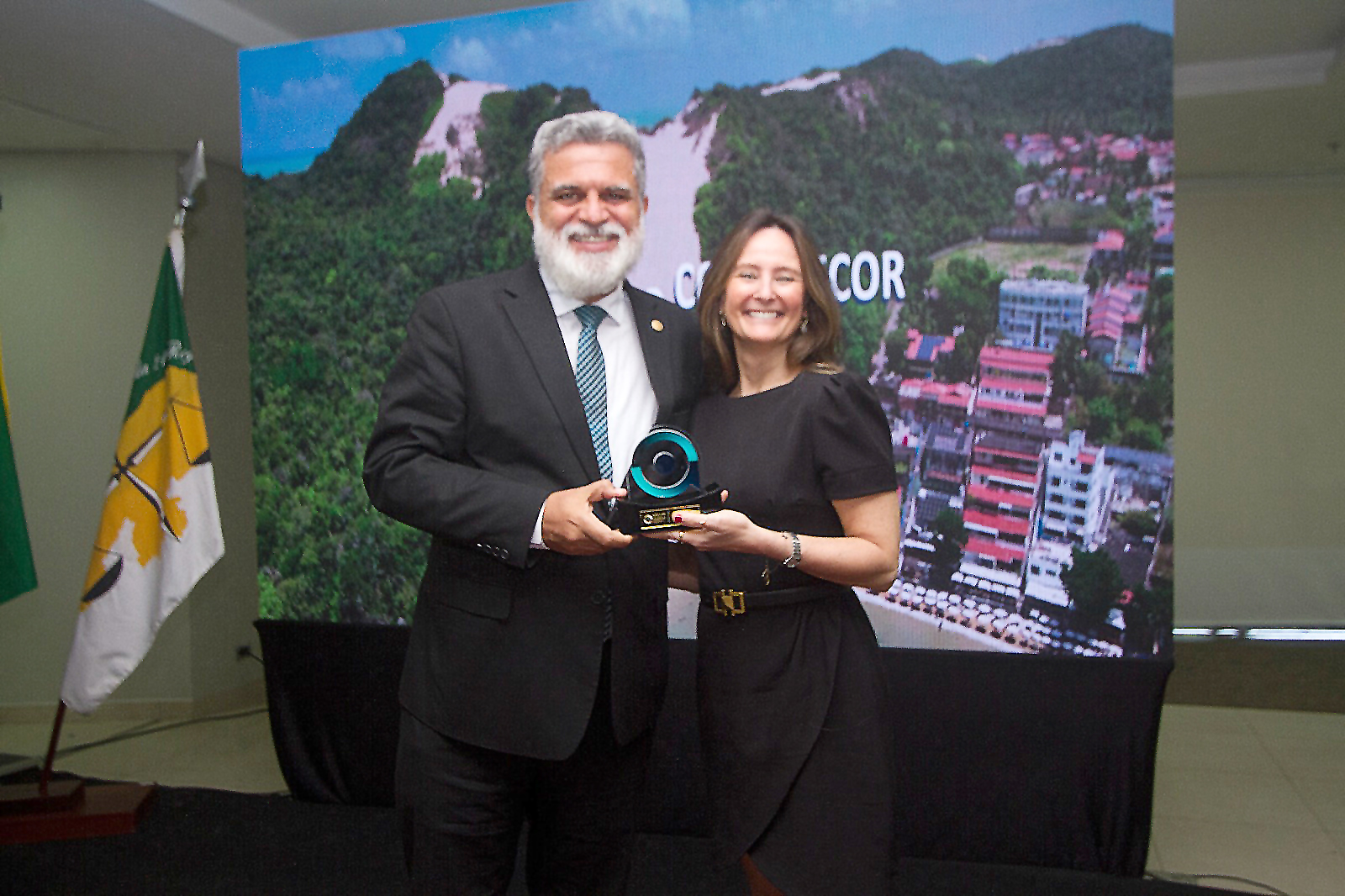 Lelio Corrêa e Luciana Xavier posam para a foto segurando um trofeu. o telão ao fundo mostra o logo do coleprecor com imagens de Natal.