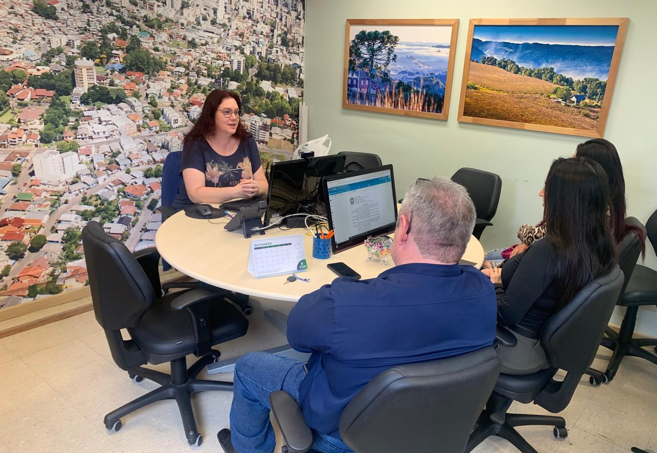 Foto de uma audiência de conciliação em Caxias do Sul. Aparecem três pessoas sentadas de costas e, em frente a elas, uma mulher conversa gesticulando. Eles estão em uma mesa redonda. Na parede de fundo aparece a foto aérea da cidade.
