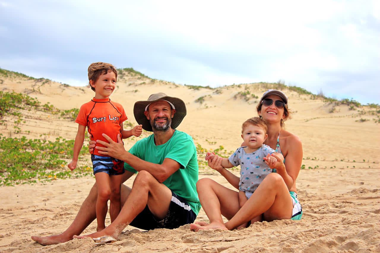 A família na praia: Isabel, Marcel, Heitor e Thales. Os pais estão sentados na areia e as crianças estão em pé.