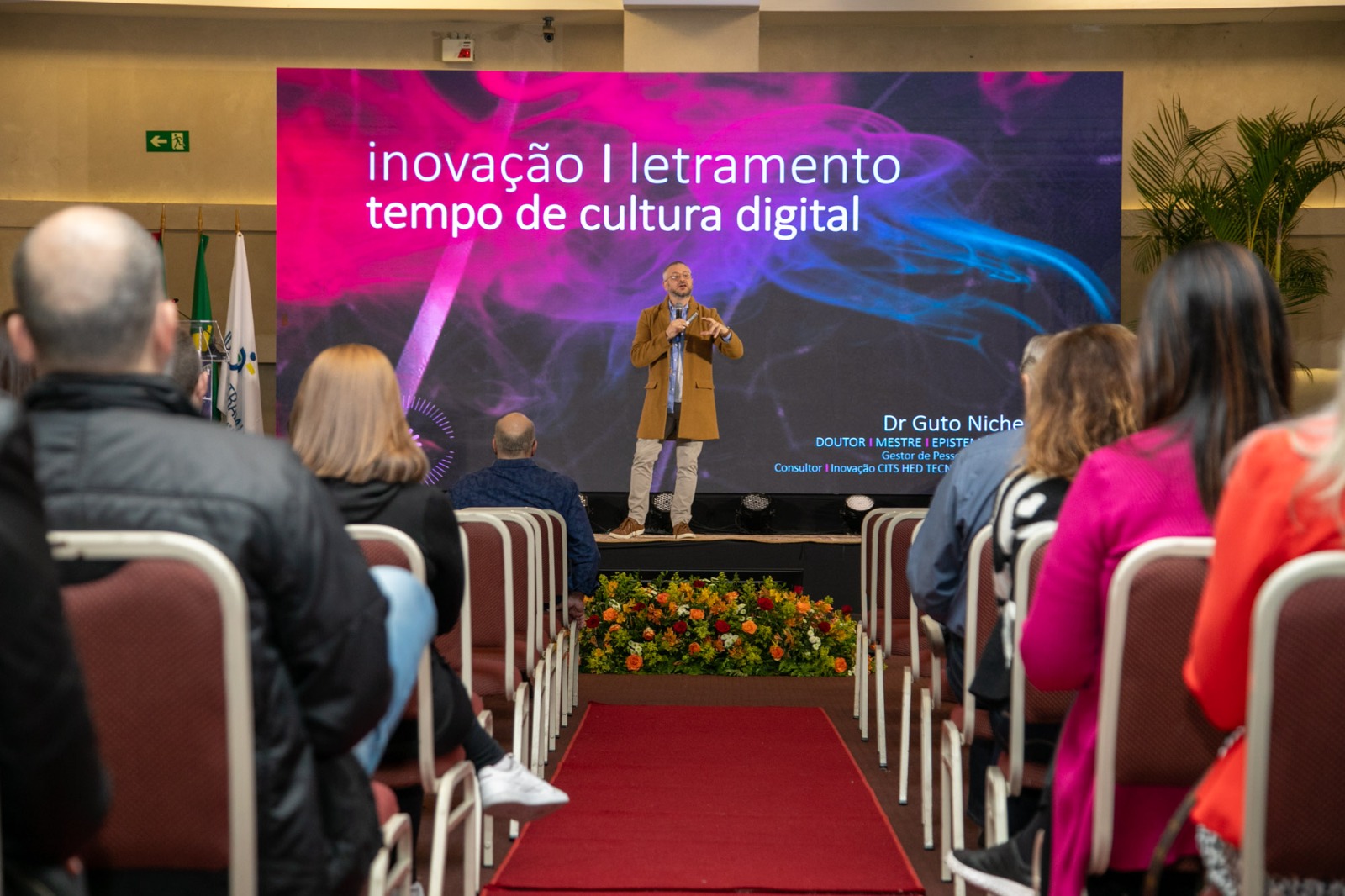 Palestrante Guto Niche falando em pé, no palco. Aparece parte do público, sentado.