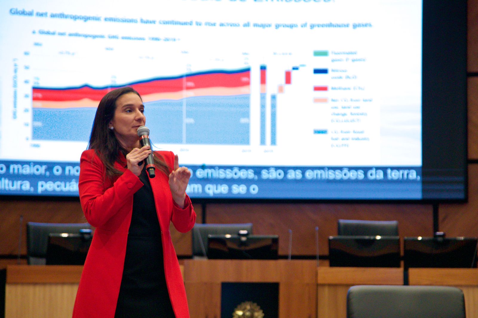 Juíza Raquela aparece falando ao microfone com um telão ao fundo. Ela é branca, tem cabelo preto escuro, e usa um blazer vermelho.