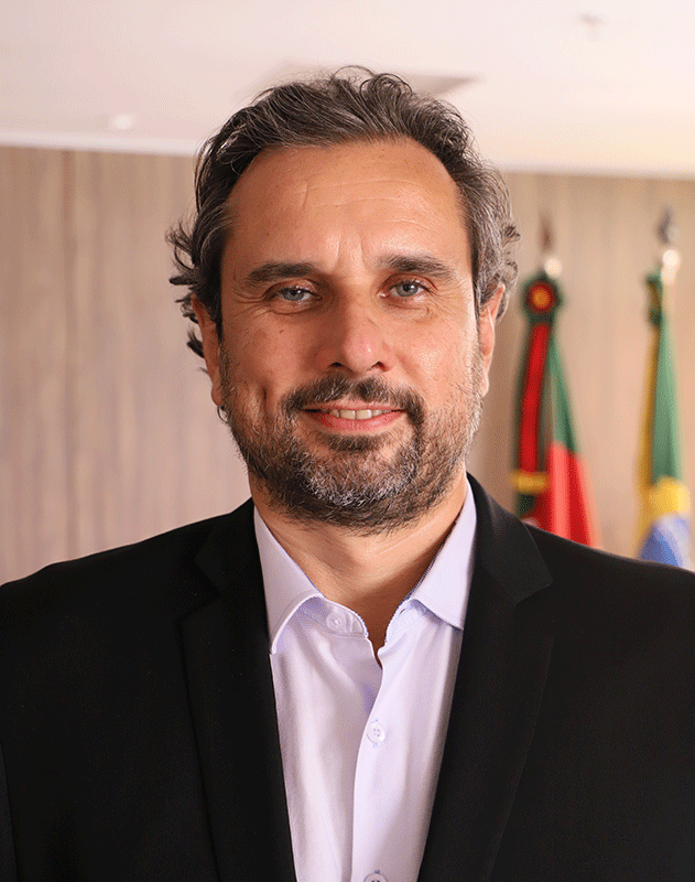 Adolfo tem cabelos e barba grisalhos, olhos claros e pele branca. Veste blazer escuro e camisa clara. Está no ambiente com bandeiras solenes ao fundo.