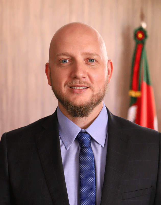 Retrato do servidor André Luiz Daiprai. Homem de pele branca, calvo. Usa barba um pouco mais comprida no cavanhaque. Os pelos são de cor castanho claro. Ele tem os olhos azuis. Usa terno escuro, camisa lilás e gravata azul com um padrão pontilhado branco