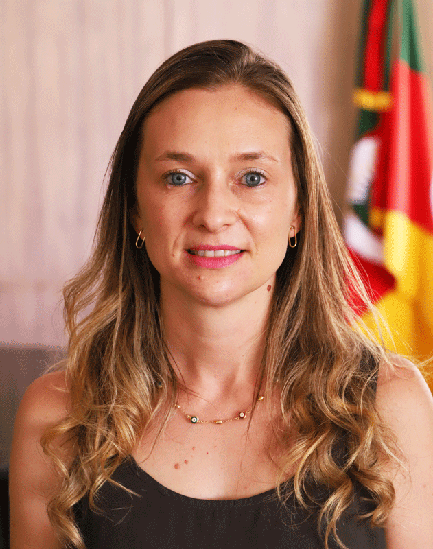 Retrato da servidora Carolina Litvin. Ela tem a pele e os olhos claros. Seu cabelo é loiro, comprido e ondulado da linha dos ombros para baixo. Usa uma blusa preta de mangas cavadas, brincos e colar dourados e bem discretos. Está maquiada com um batom cor de rosa