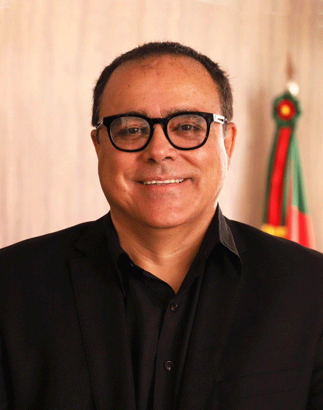 Retrato de Geraldo Teixeira. Ele tem a pele clara, cabelo e olhos castanhos. O cabelo é bem curto. Usa óculos de armação preta e veste um blazer e uma camisa também pretos.