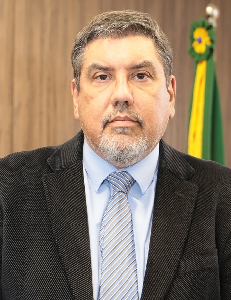 Retrato de Aldo Jardim. Homem branco de cabelos grisalhos, veste terno e gravata. Ao fundo, bandeira cerimonial do Brasil