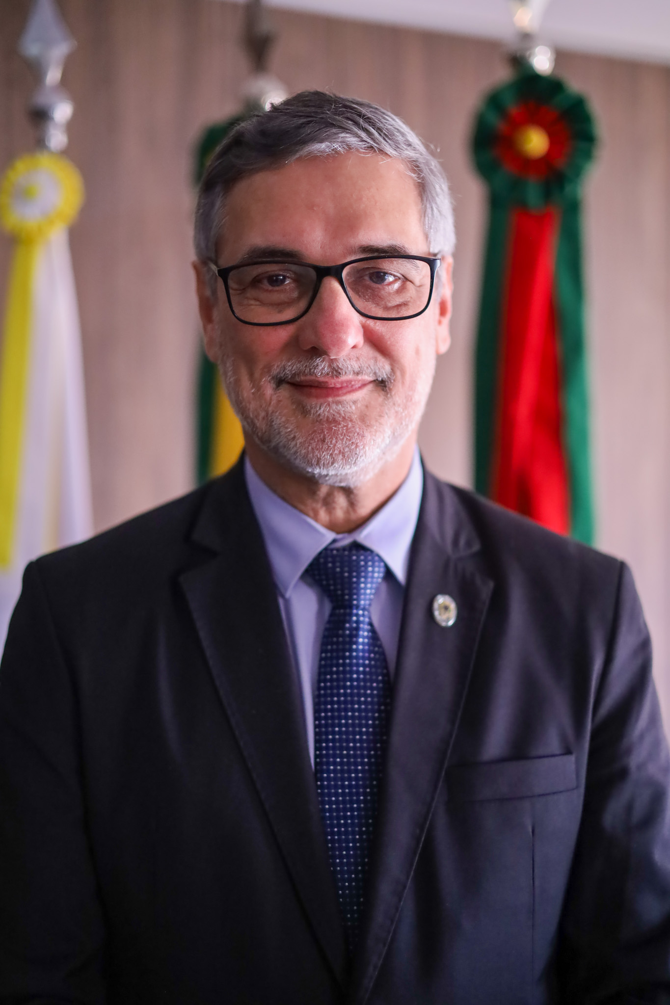 Retrato de João Luiz Peixoto. Ele tem cabelo e barba grisalha, usa óculos de armação escura e sorri. Ao fundo, bandeiras solenes.