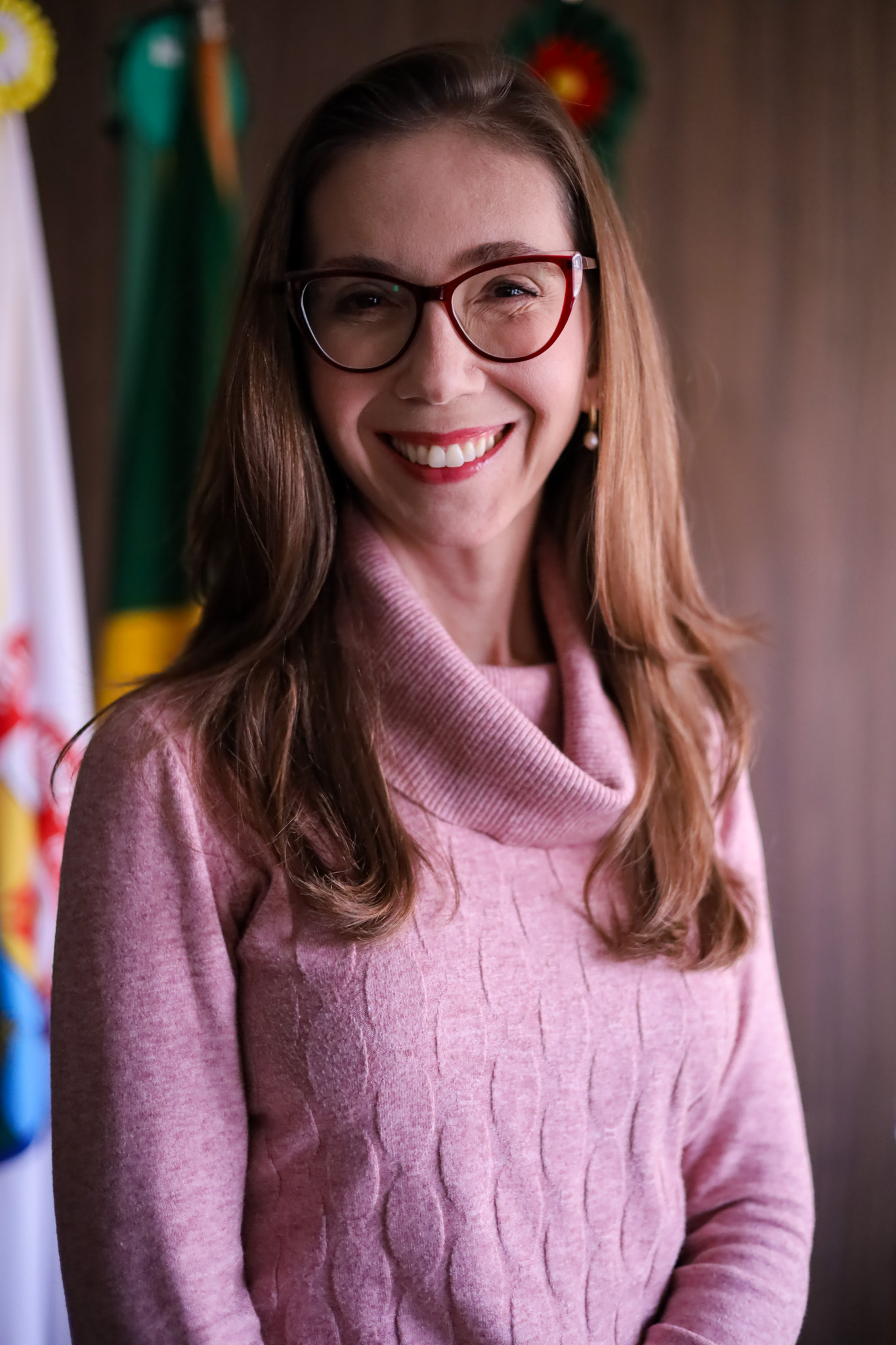 Retrato de Paula Rosa. Mulher de cabelos castanhos claros compridos, pele branca, sorri e veste óculos de aros bordô e blusa rosa. Ao fundo, bandeiras solenes e parede marrom acinzentada.