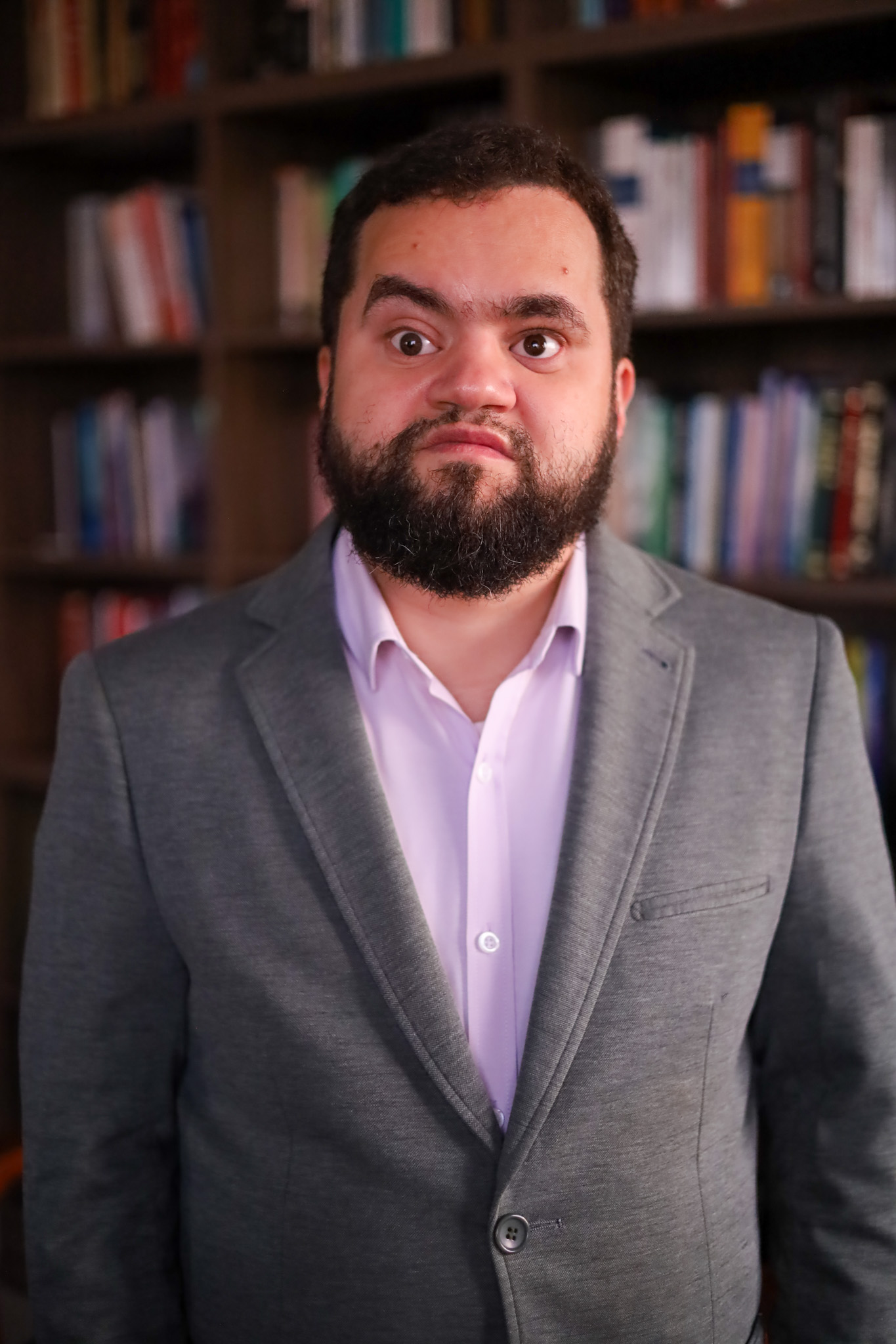 Retrato de Juliano Machado. Juliano é homem branco de cabelos e barba pretos, veste blazer cinza e camisa clara. 