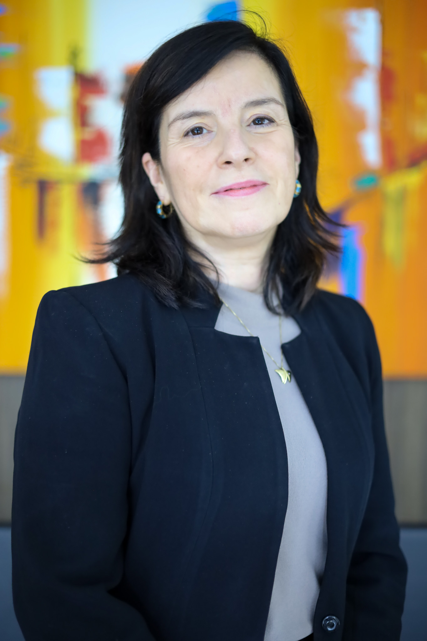 Retrato de Enilda Andrade. Mulher branca de cabelos pretos de médio comprimento, sorri. Veste blazer preto com blusa cinza claro e corrente dourada com pingente e brincos azuis e dourados.