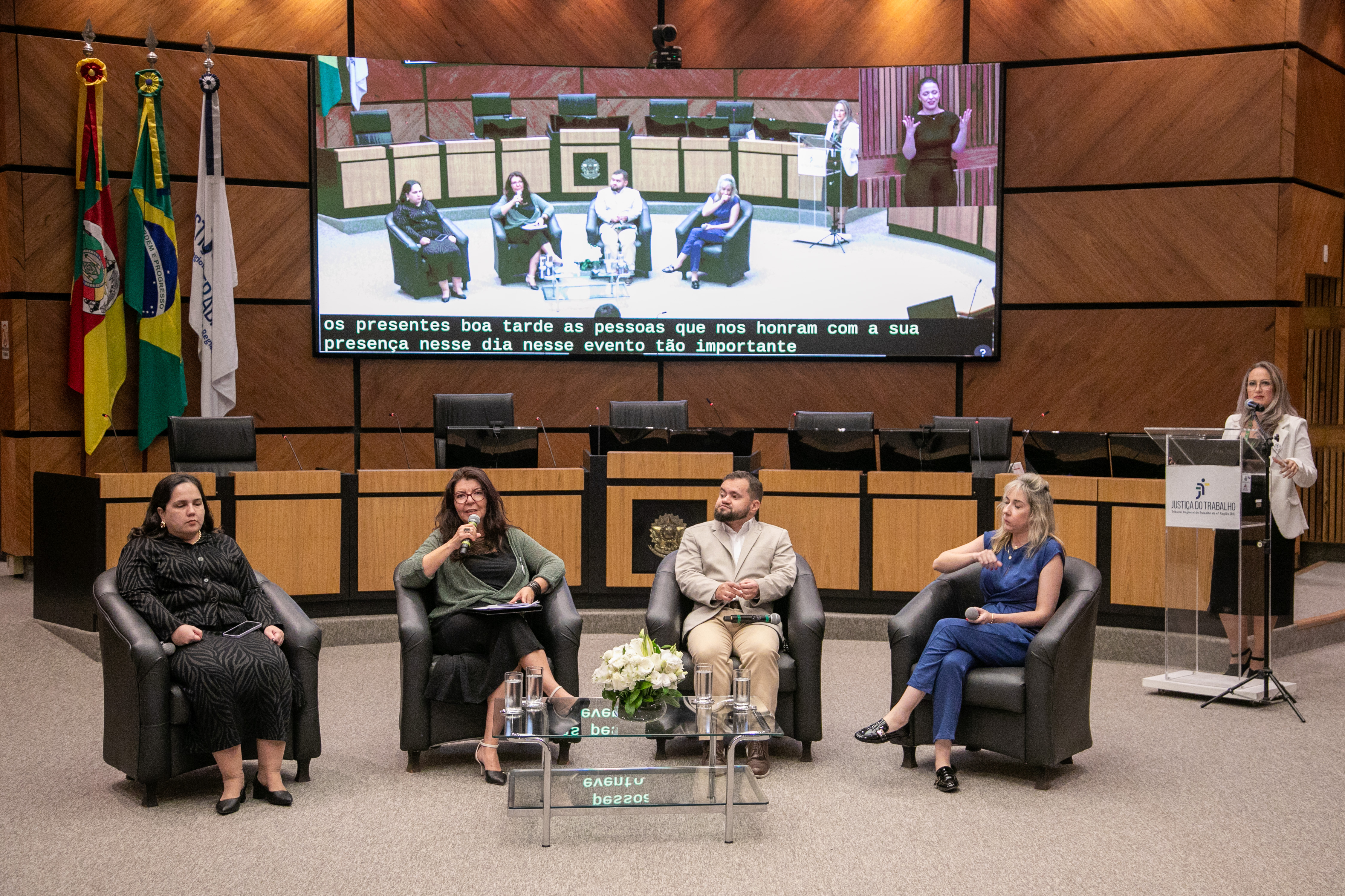 Painelistas do evento Quatro palestrantes estão sentados em poltronas pretas, dispostos em semicírculo. À esquerda, Juliana está de vestido preto com estampa animal print observa atenta. Ao lado dela, Lúcia, de óculos e casaquinho verde, segura o microfone e fala ao público. À direita, Juliano, que veste um terno bege e sapatos marrons, escuta atento. Na ponta, Renata, que está de macacão azul, segura um microfone no colo, acompanhando a fala. À frente, mesa de vidro com flores brancas e copos de água.