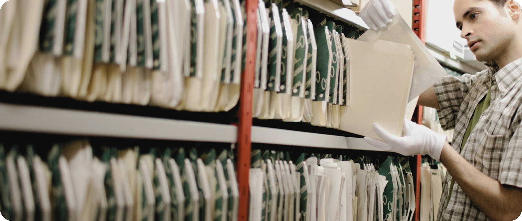 Imagem de uma pessoa com luvas manuseando uma pasta com documentos em frente a uma estante cheia de pastas.