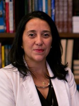 Maria Teresa é parda, de cabelo preto e liso. Sorri para a foto. Ao fundo há uma biblioteca. 