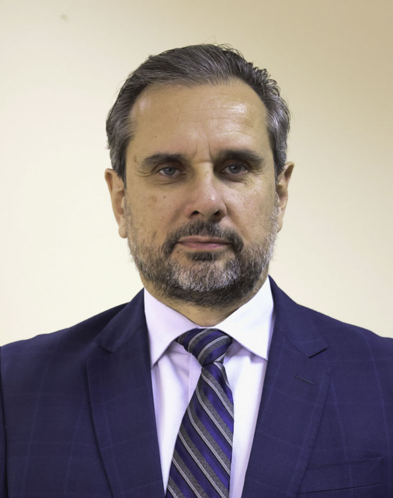 Retrato de Adolfo. Ele é branco, barba e cabelo grisalhos e de olhos claro. Veste blazer azul xadrez,. camisa social branca e gravata listrada em azul, branco e cinza. 