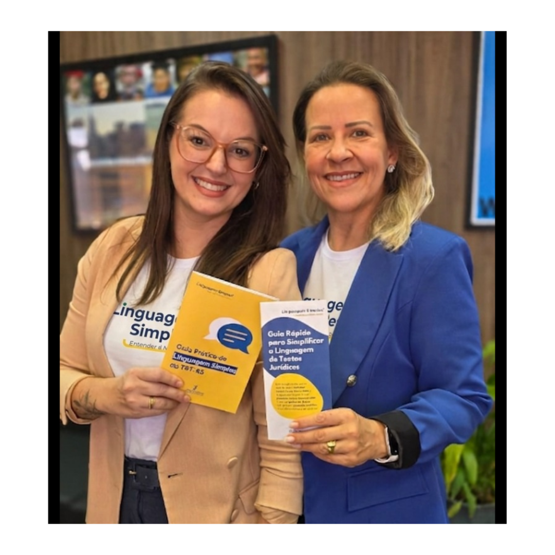 Duas mulheres sorridentes posam juntas para uma foto. Uma delas usa blazer bege e a outra blazer azul. Elas seguram duas publicações impressas: um livreto amarelo com o título "Guia Prático da Linguagem Simples no TRE-RS" e um panfleto azul intitulado "Guia Rápido para Simplificar a Linguagem de Textos Jurídicos". Ao fundo, vê-se uma televisão e algumas plantas. Duas mulheres sorridentes posam juntas para uma foto. Uma delas usa blazer bege e a outra blazer azul. Elas seguram duas publicações impressas: um livreto amarelo com o título "Guia Prático da Linguagem Simples no TRE-RS" e um panfleto azul intitulado "Guia Rápido para Simplificar a Linguagem de Textos Jurídicos". Ao fundo, vê-se uma televisão e algumas plantas.