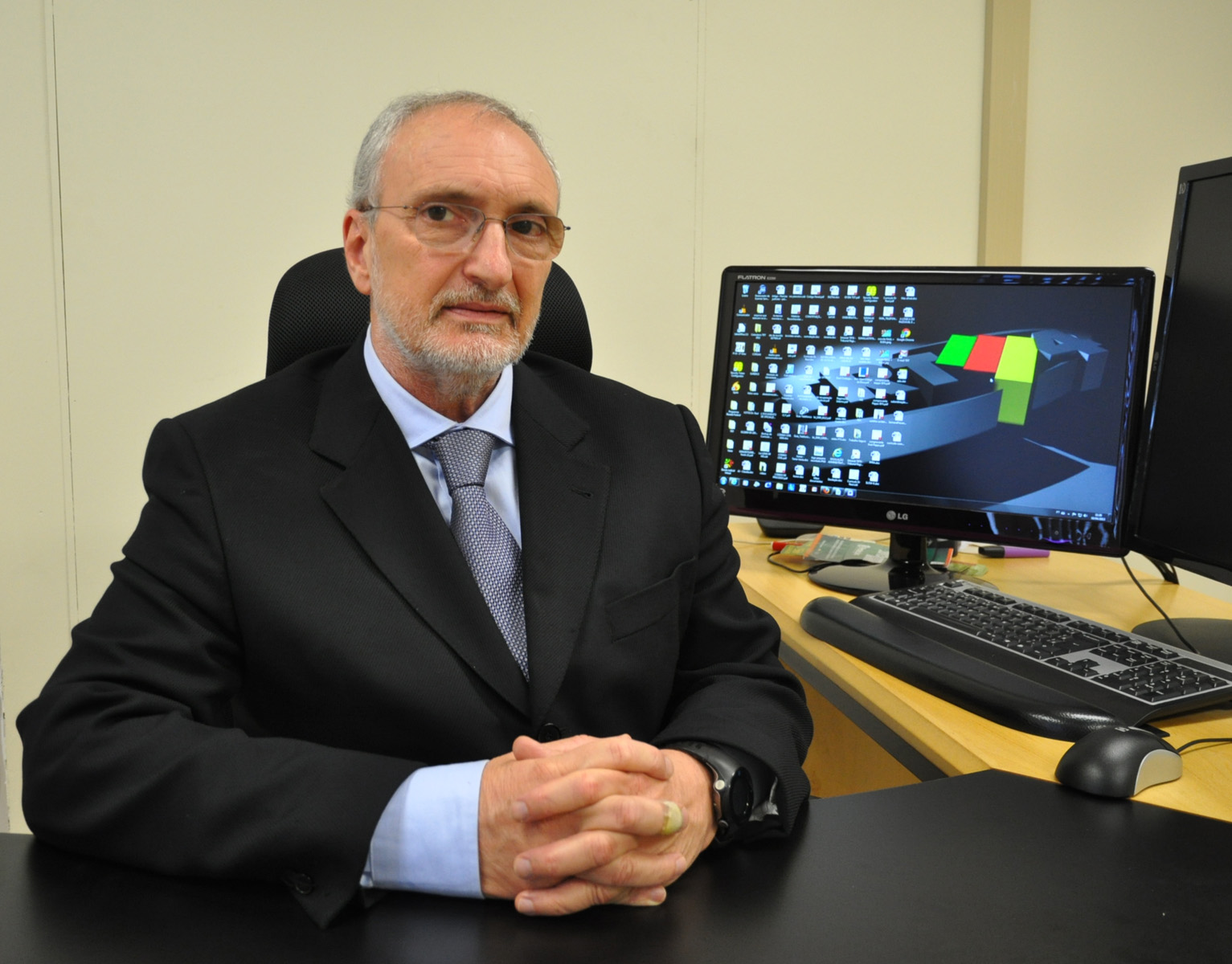 Foto do desembargador em seu gabinete. Homem branco, de barba grisalha curta, veste óculos e repousa as mãos sobre a mesa.